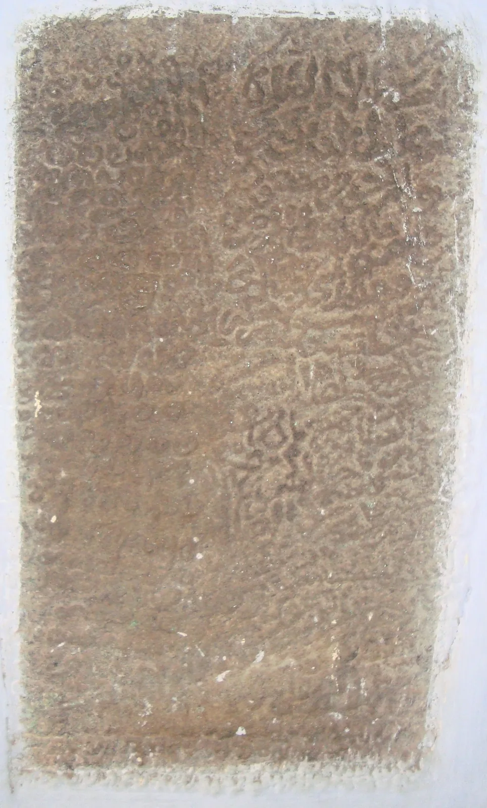 The Muccunti mosque inscription: a worn brown granite slab filling the frame, its lettering standing in low raised relief, the surrounding whitewash carried over the edges of the stone.