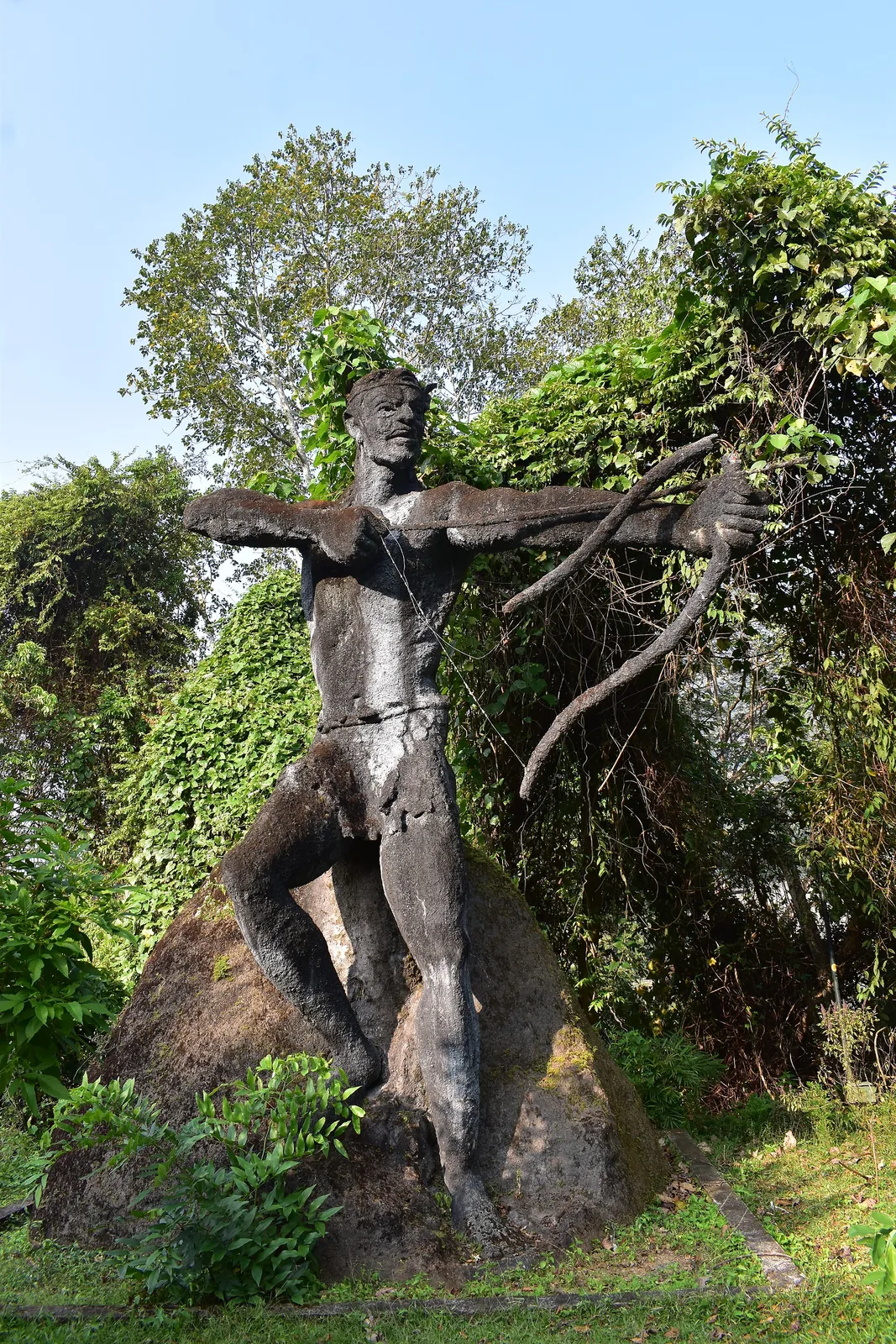 A large weathered dark stone sculpture of a lean, near-naked archer at full draw, standing astride a rock outcrop in the Thenmala sculpture garden, a thin wire standing in for the bowstring and green creeper-covered trees behind.