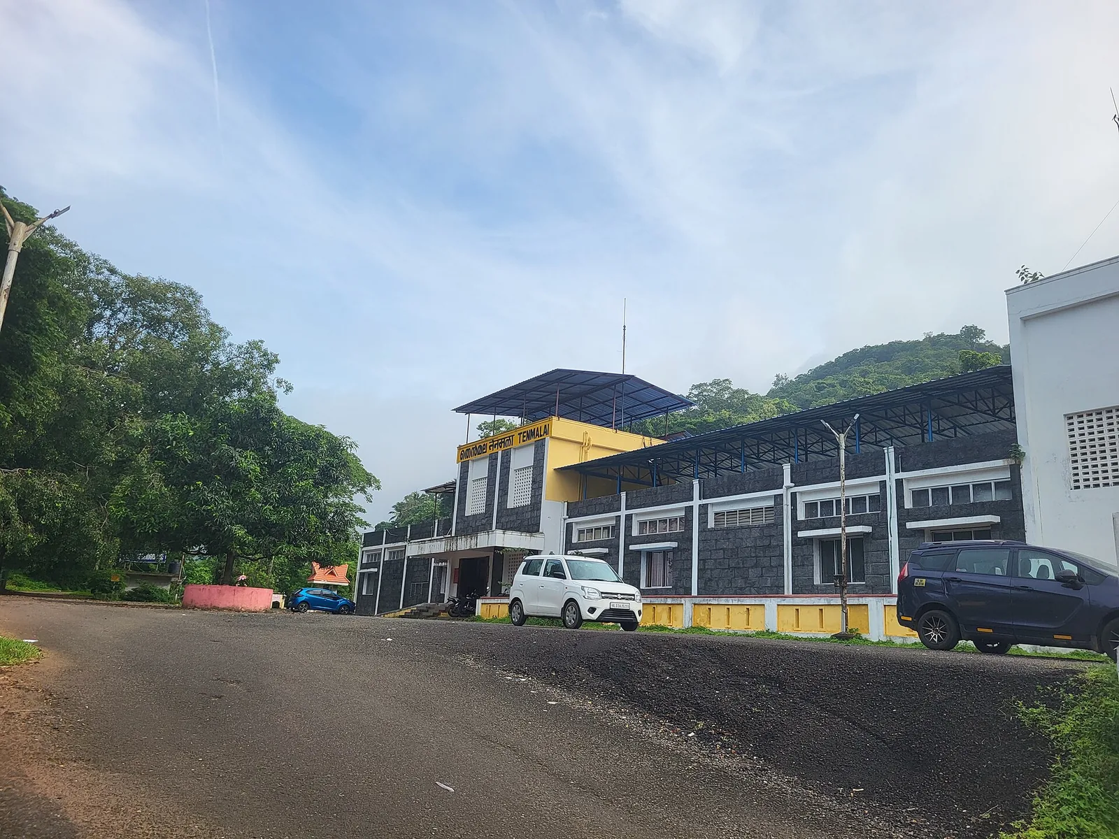 Tenmalai railway station on a bright morning: a low grey stone building with a yellow upper block carrying the nameboard, a blue steel platform canopy running to the right, forested hills behind and three cars parked on an otherwise empty forecourt.
