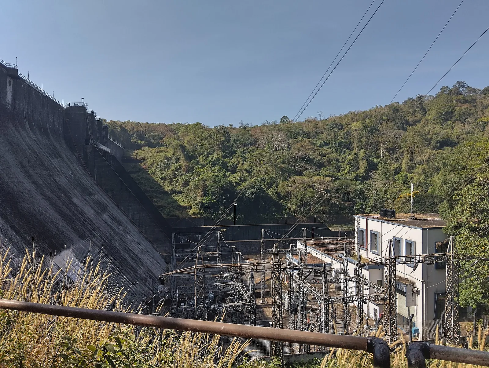 The KSEB switchyard below the Thenmala dam in dry-season light — lattice gantries, insulator strings and transformers in the foreground, a white two-storey powerhouse block behind them, and the steeply raking grey masonry face of the dam filling the left of the frame.