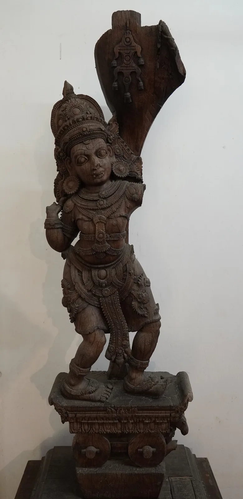 A tall carved figure of Shiva as Thripuranthaka, holding a bow, ornamented and crowned, displayed in a museum gallery