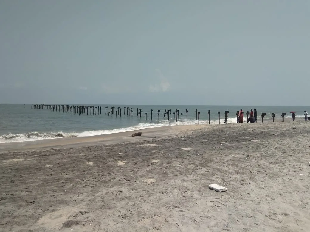 The rows of rusted iron stumps of the old sea piers running out from Alappuzha beach into a flat grey sea, with people walking on the sand