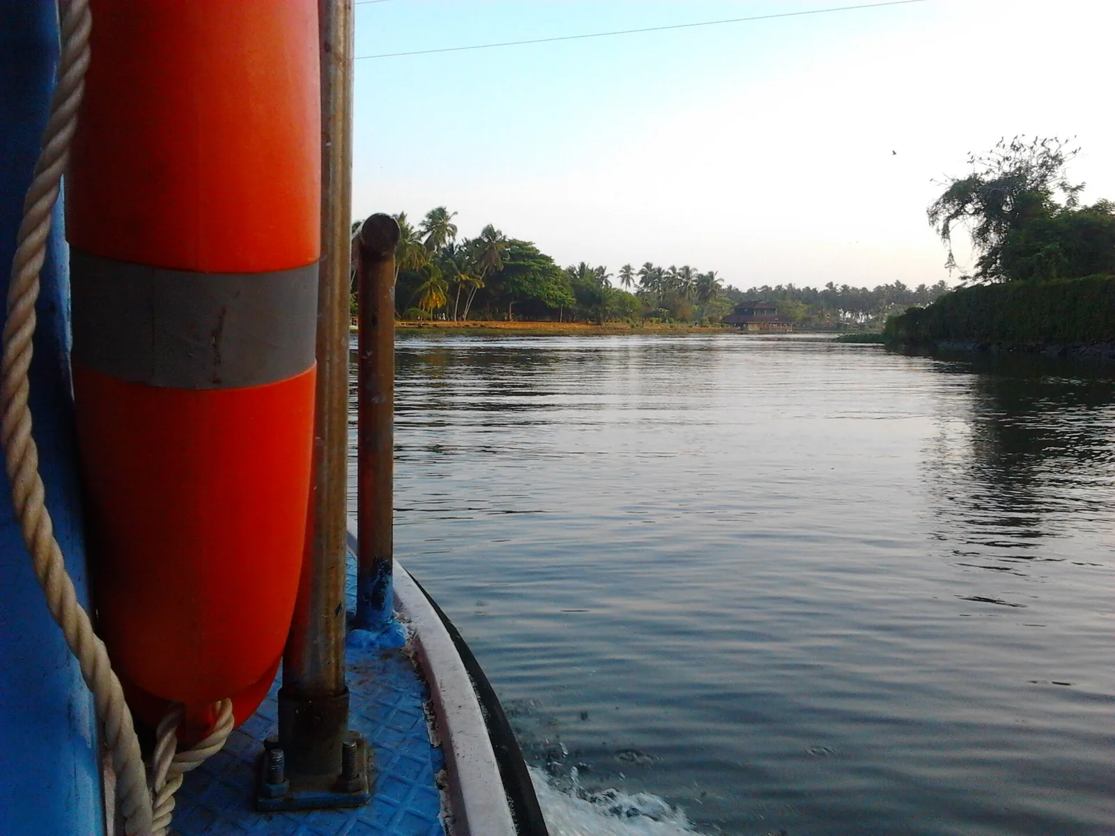The bow of a boat with an orange life buoy, running on the Veli backwater past wooded banks
