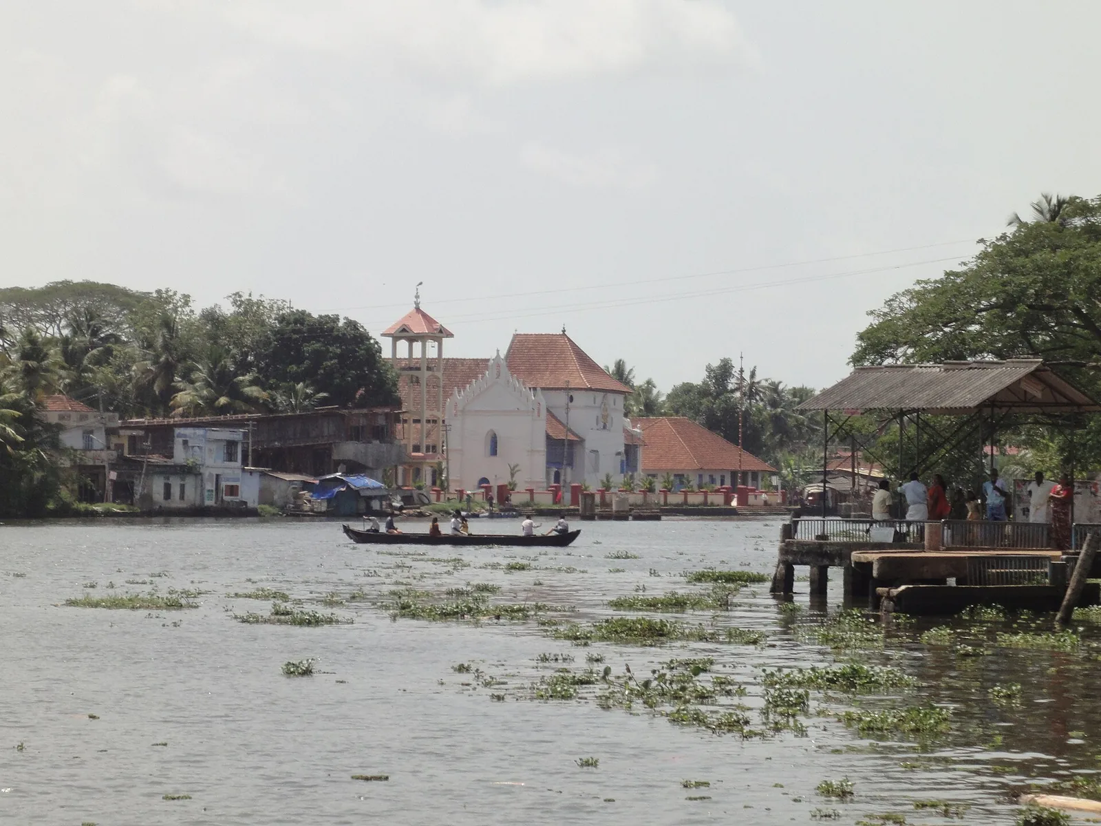 The Pamba river at Champakulam, its surface strewn with water hyacinth, a country canoe crossing with passengers aboard, and a roofed ferry jetty with people waiting on the right bank