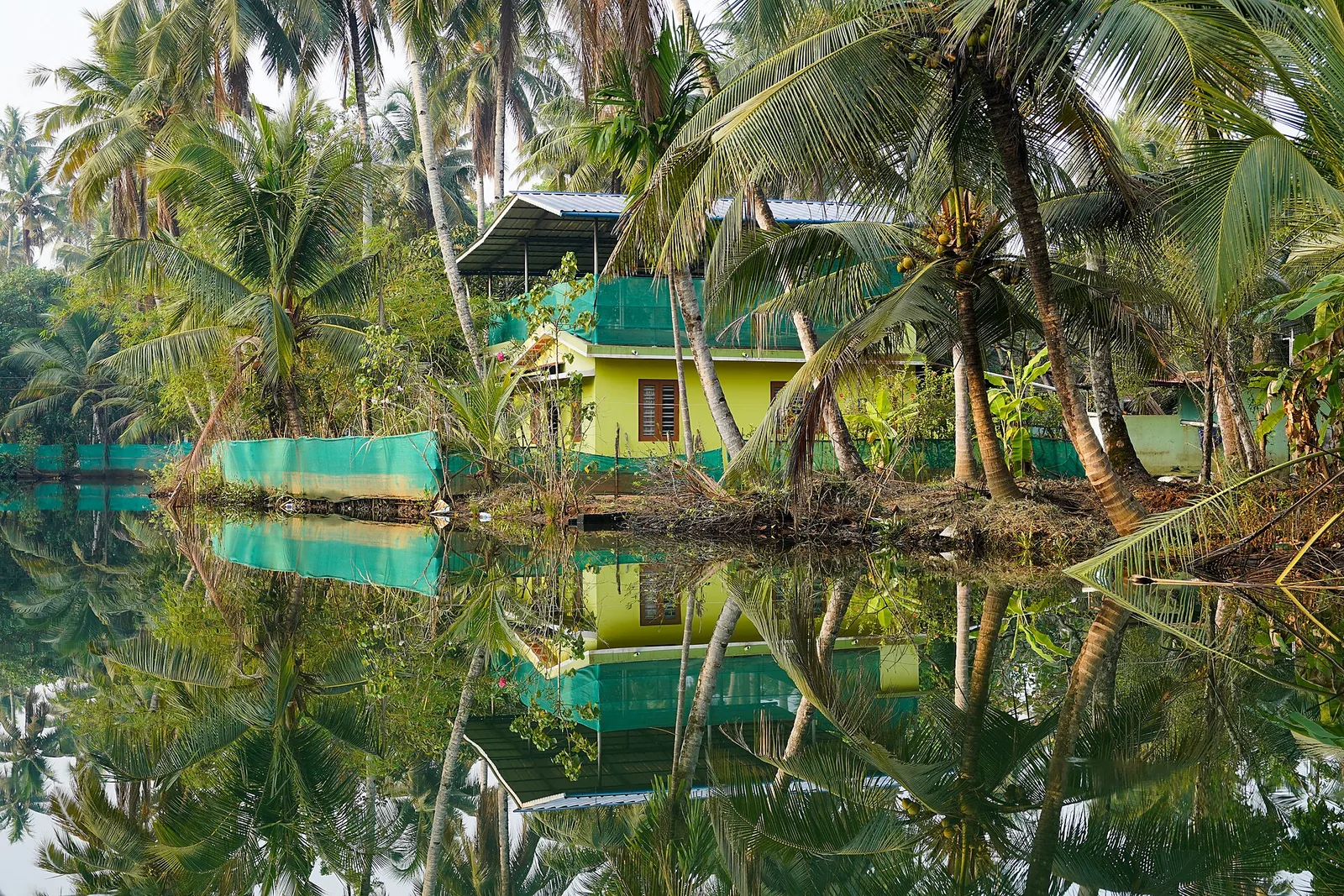A yellow-and-green house on Munroe Island standing among coconut palms on the bank of a still canal, its whole reflection held perfectly in the water, with an open unwalled extra storey under the roof.