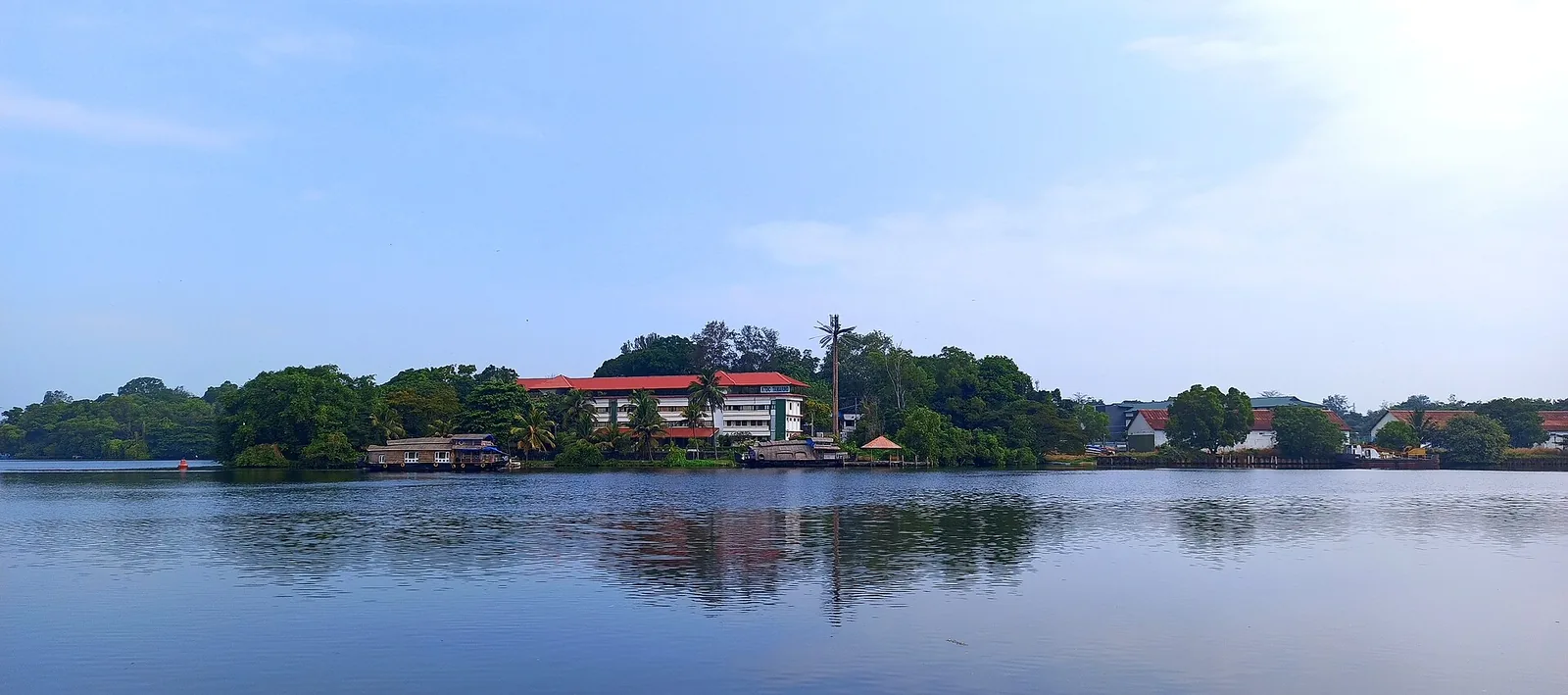 A wide sheet of still blue water on Ashtamudi under a pale sky, with a built-up far shore of trees, small waterside sheds and a long red-roofed multi-storey building reflected on the surface.