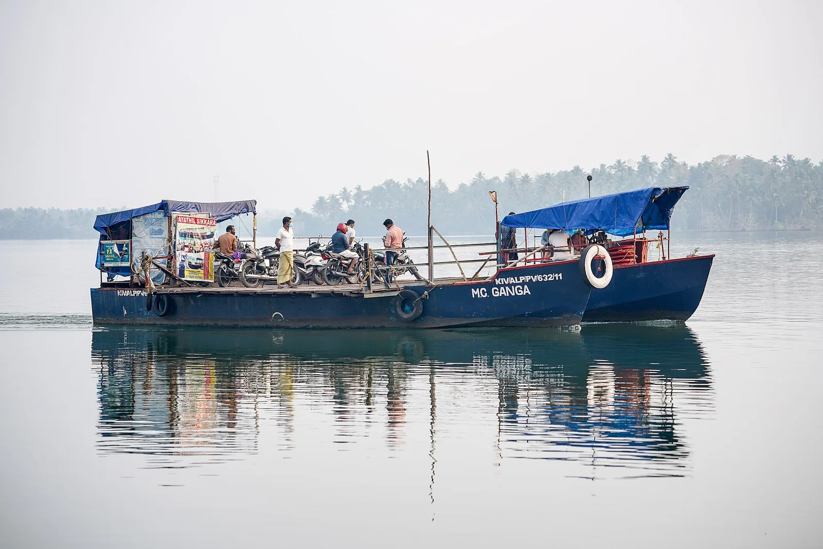 A blue-hulled flat-decked ferry named MC Ganga crossing calm grey-green water on Ashtamudi, about a dozen motorcycles parked across its open deck under a blue tarpaulin canopy, a few passengers standing among them and a low palm-covered shore in the haze behind.