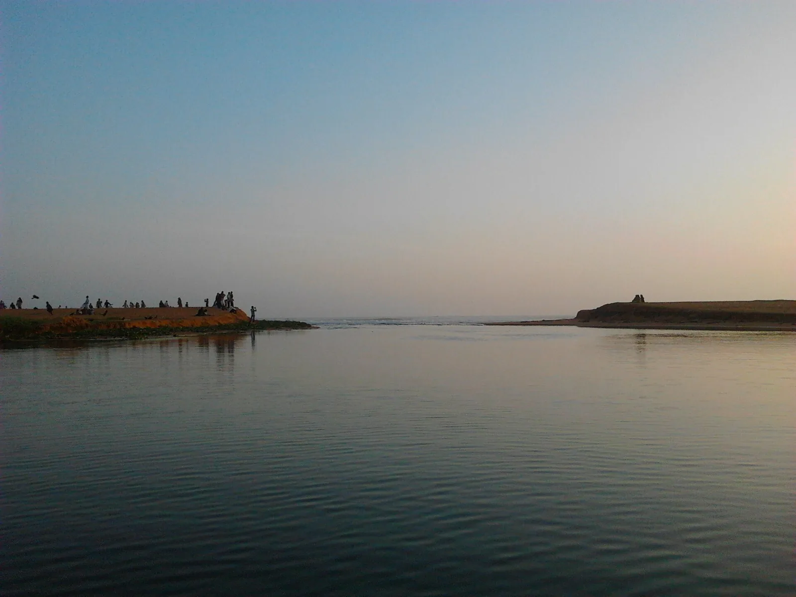 The opening where Veli lagoon meets the Arabian Sea between two low sand banks, with people standing on the bar at dusk