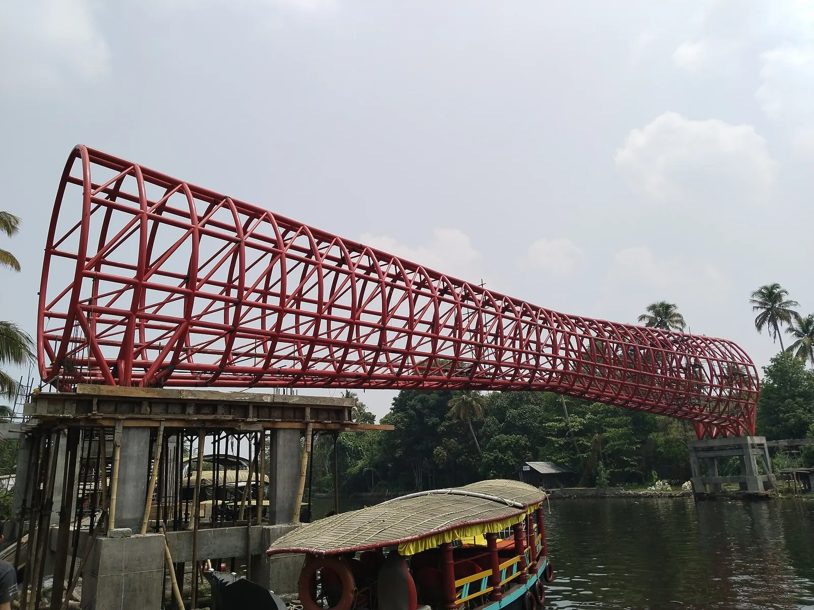 A red steel lattice footbridge over water at Punnamada, tapering from a wide flared mouth to a narrow tail like a snake boat