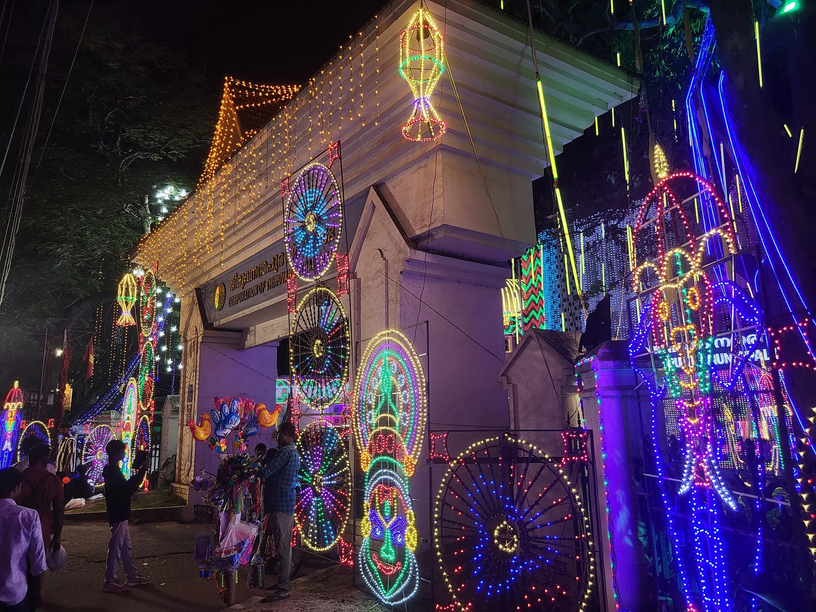The gateway to the Kanakakunnu grounds at night during Onam, hung with illuminated wheels, lamps and figures in coloured lights