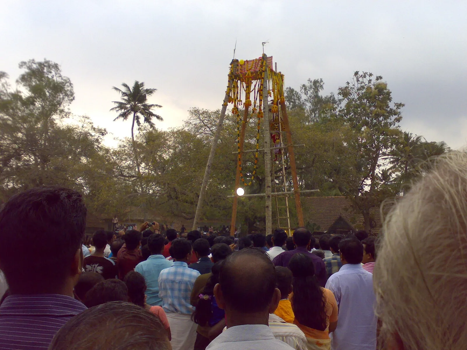 A tall garlanded wooden festival structure rising above a dense crowd at the Kaliyoot, Sarkaradevi Temple, Chirayinkeezhu