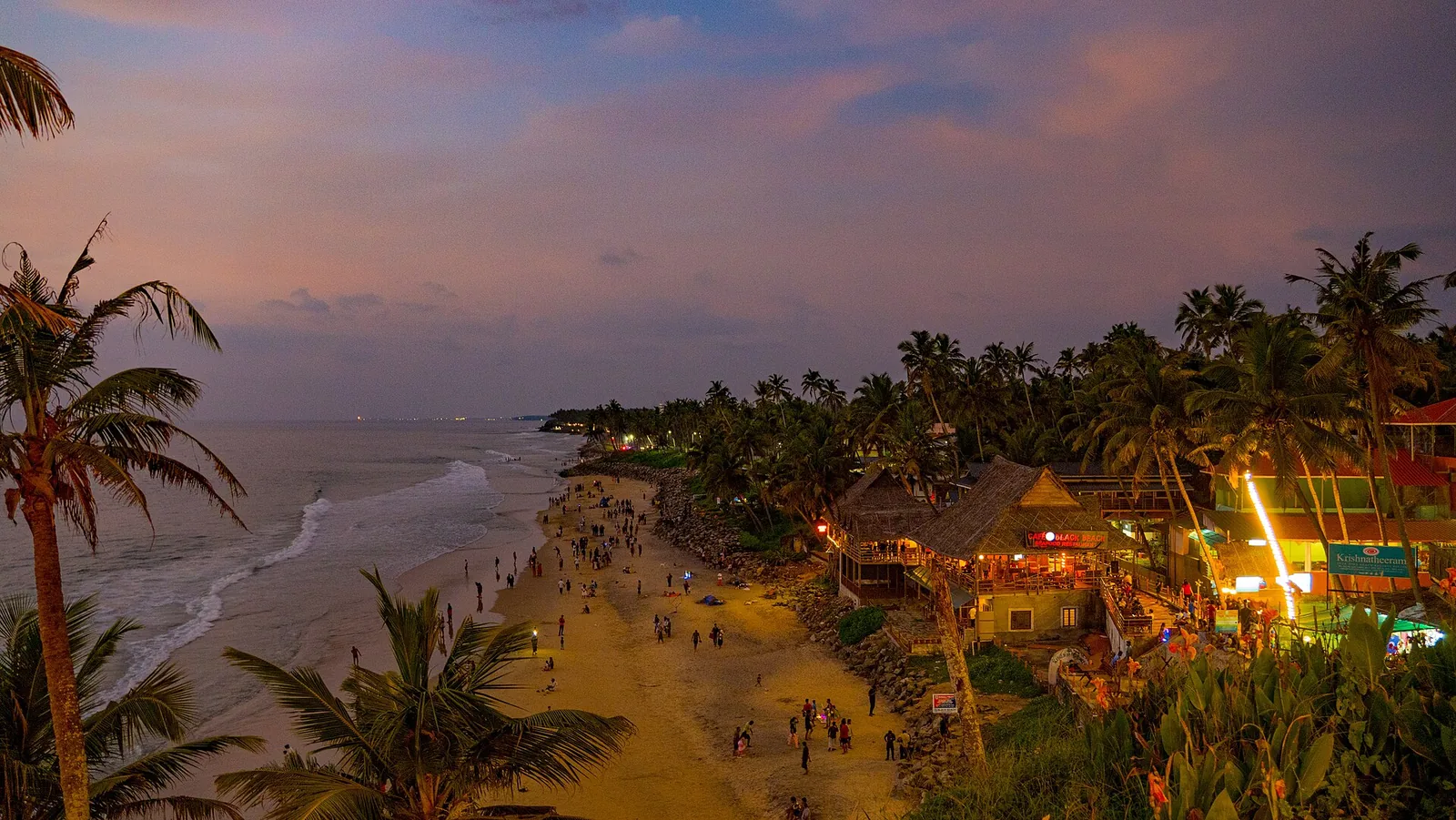 The lit café strip along the top of Varkala cliff at dusk, with rock armour piled at the base below