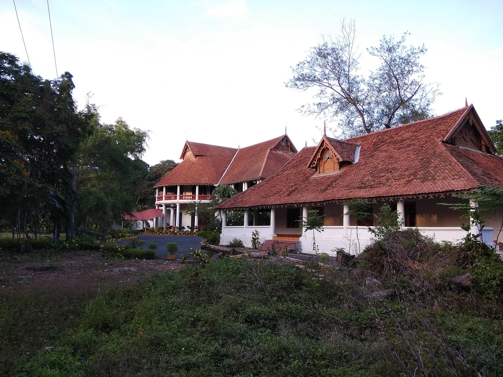 The rear of the British Residency at Asramam: a long single-storey wing with a deep verandah on white columns under a tiled hipped roof with a carved gable, the two-storey block with its red balustraded verandah standing behind it