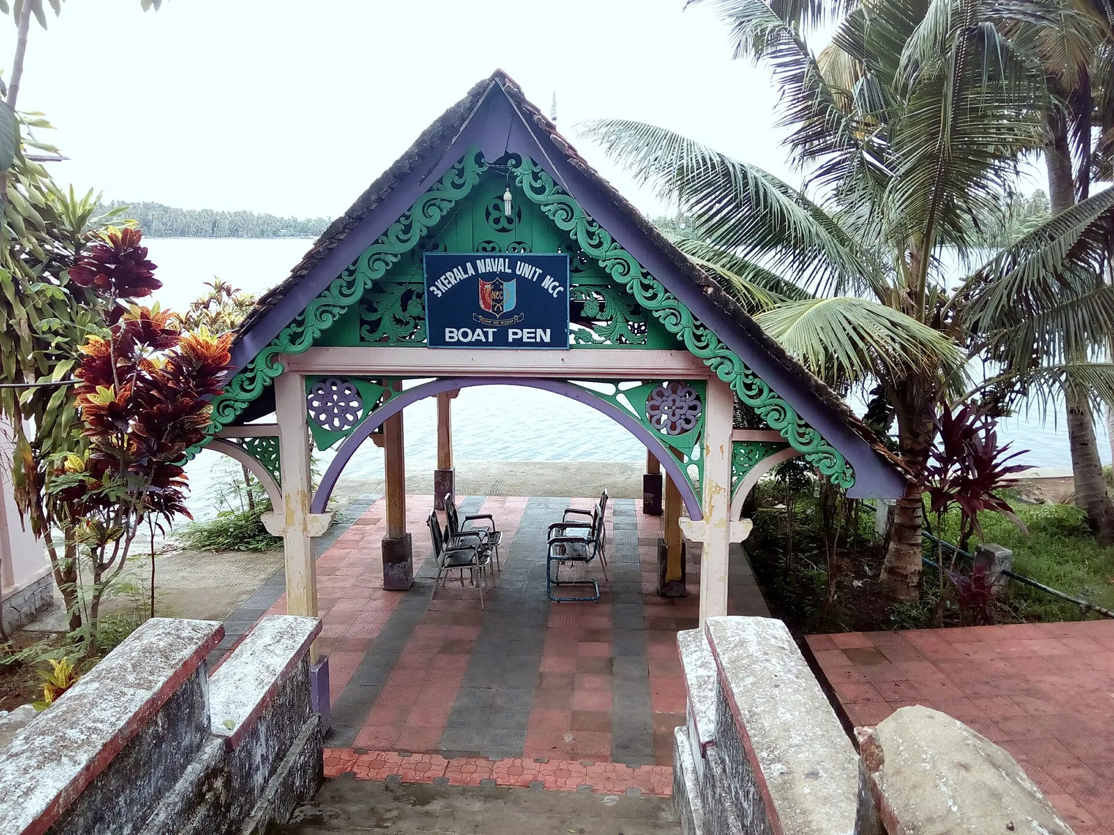 A small carved gabled pavilion painted green, purple and cream at the head of the steps down to Ashtamudi Lake at Thevally Palace, its fretwork bargeboards and pierced rosettes framing a board reading “3 Kerala Naval Unit NCC — Boat Pen”, with steel chairs facing the water.