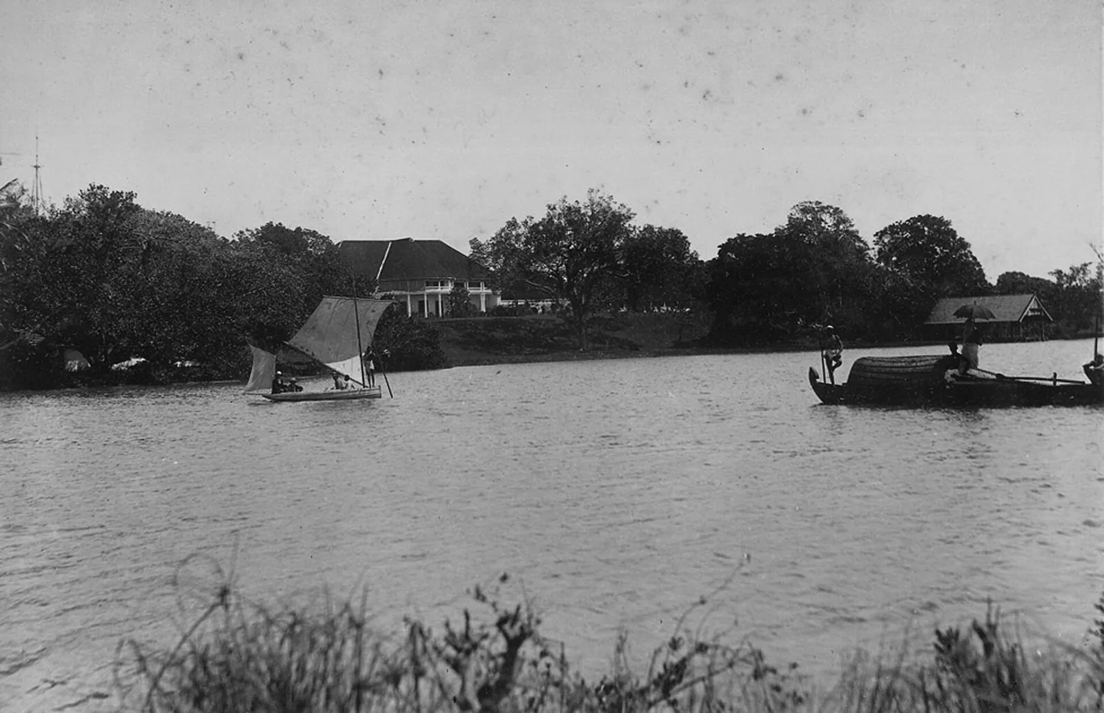 A black and white photograph from the early 1900s looking across a wide backwater to the British Residency at Kollam, a low colonnaded house with a hipped roof standing on a green rise among trees, with a small sailing boat and a covered cargo boat on the water in the foreground