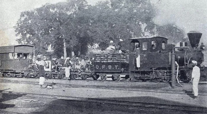 A small steam locomotive lettered C.F.S.T. with a rake of flat wagons crowded with workers and a covered caboose, standing on level track under large trees
