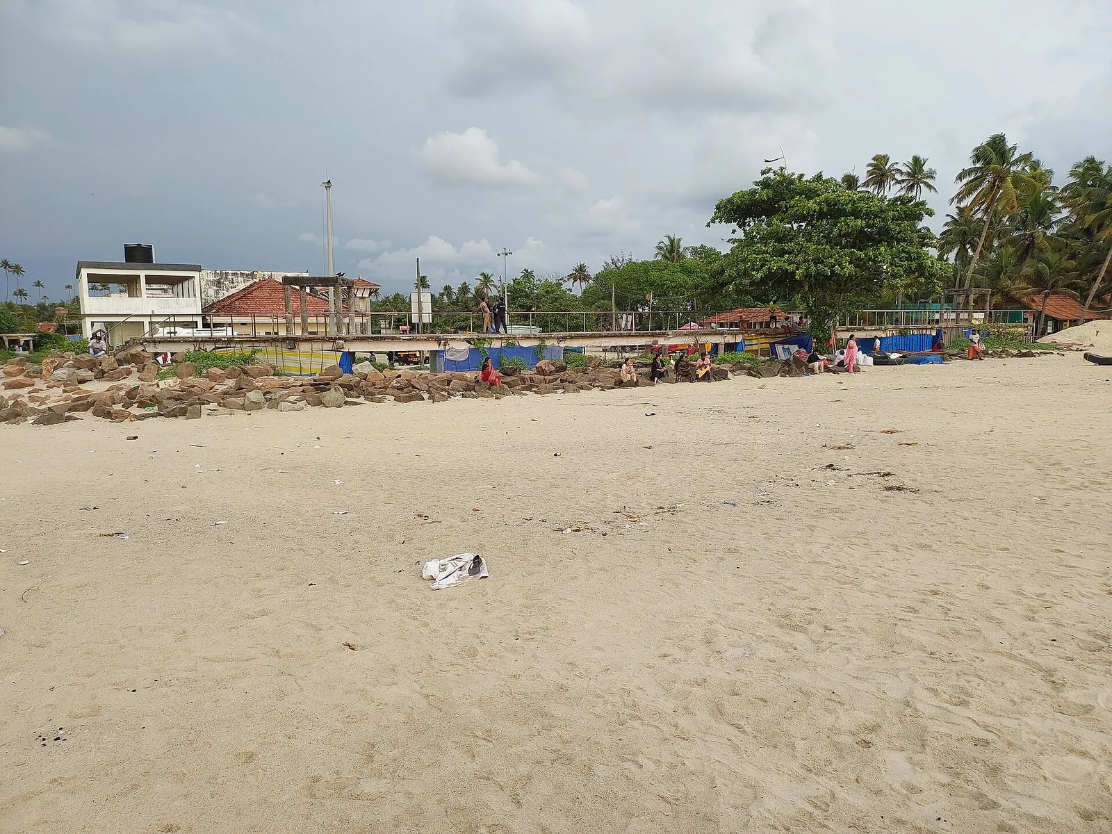 The wide pale sand of Andhakaranazhi beach with the footbridge structure and laterite boulders behind, and people sitting along the rocks