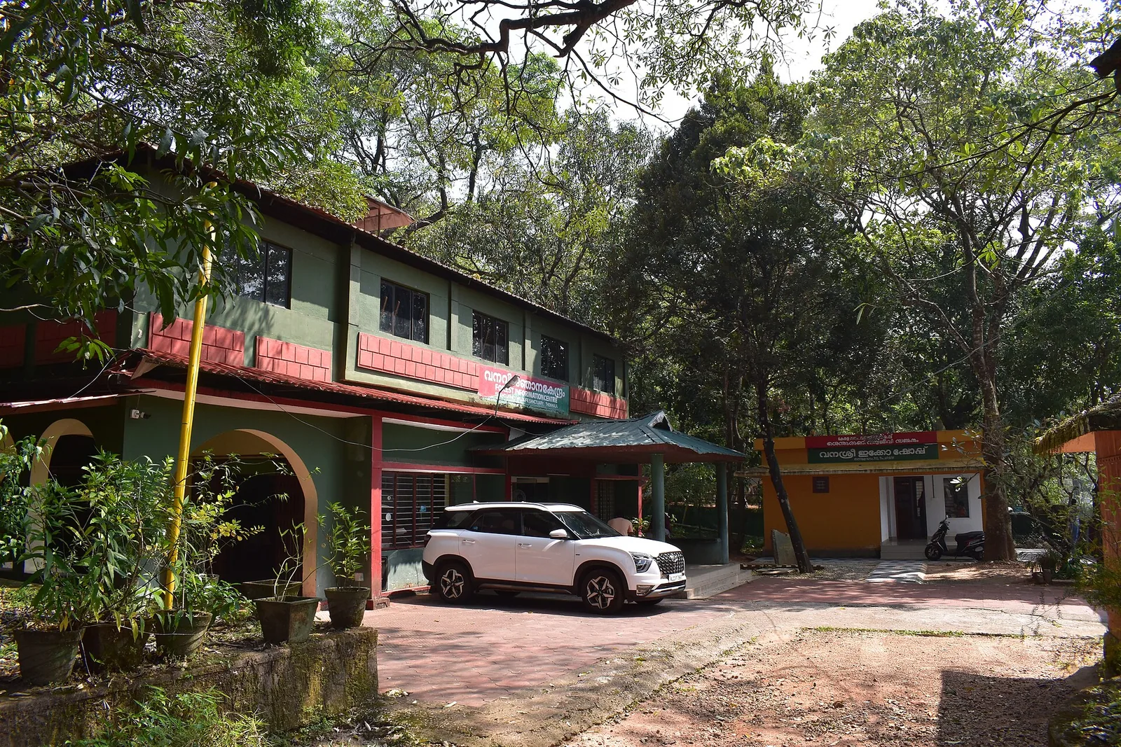 A two-storey green and dark-red Forest Department building with Malayalam signboards under a tiled roof, a car parked on the apron, and a small orange-walled eco shop beside it under tall trees.