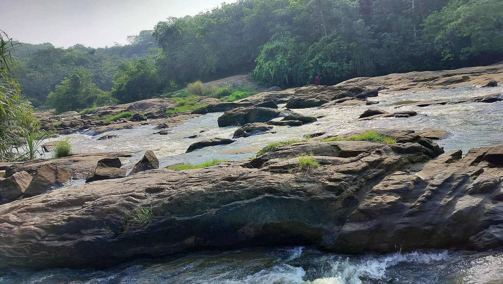 Long fluted ledges of grey rock stepping down into the river, with white water braiding between them and a single figure in red standing on the far bank for scale