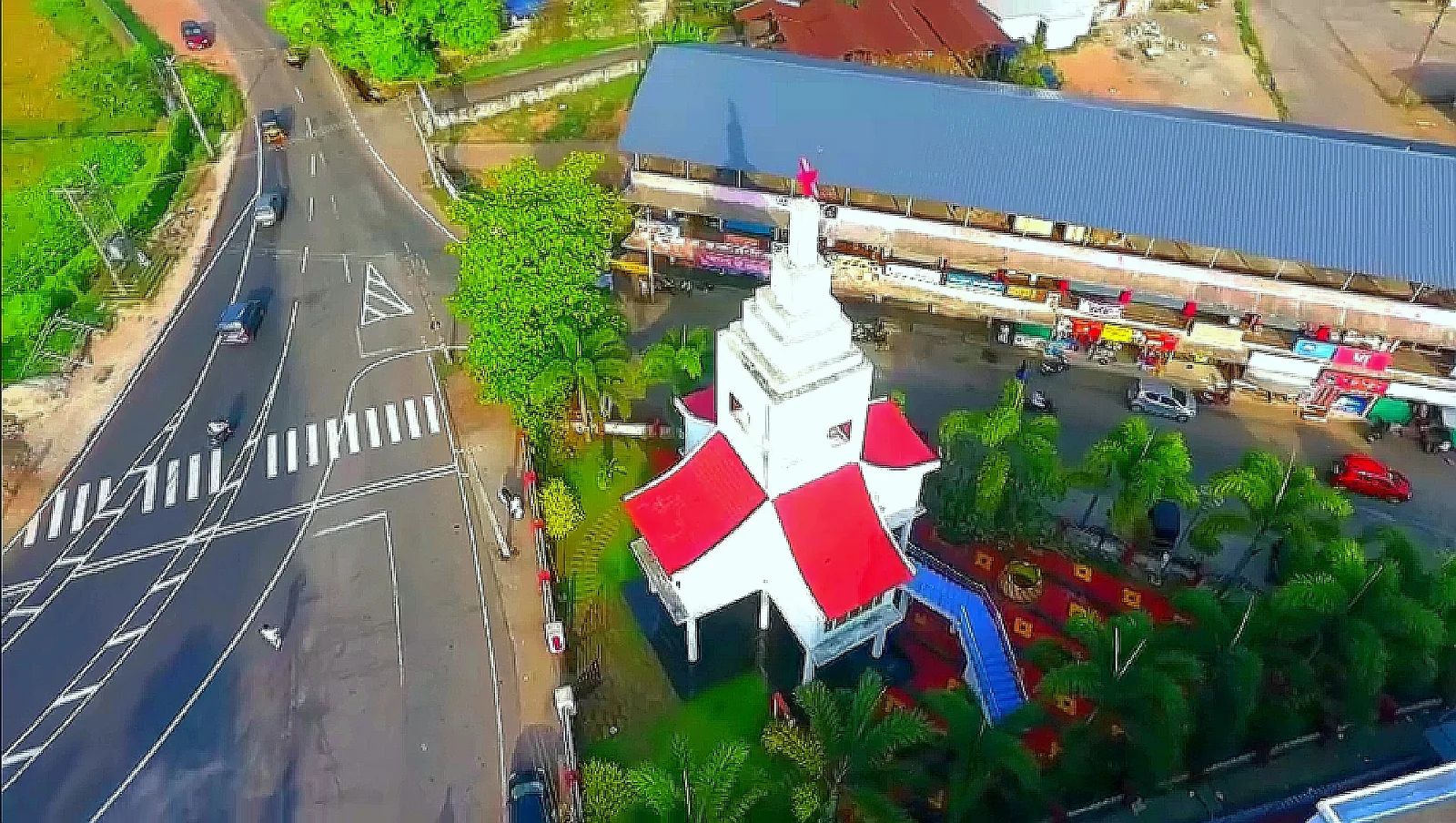 Aerial view of a white tiered memorial tower with red and white pitched roofs and a red star at its top, standing in a small landscaped plot at a road junction beside a long market shed, with traffic on the road alongside.