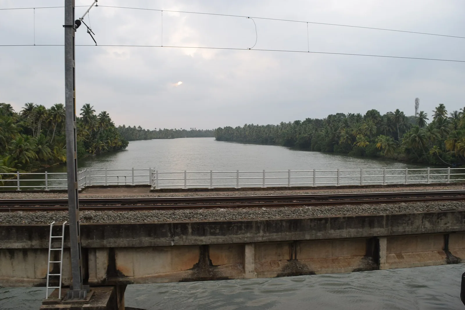 The Kallada river seen over the parapet of a railway bridge, a broad calm reach running away between dense coconut palms under an overcast sky, with overhead electrification wires crossing the frame.