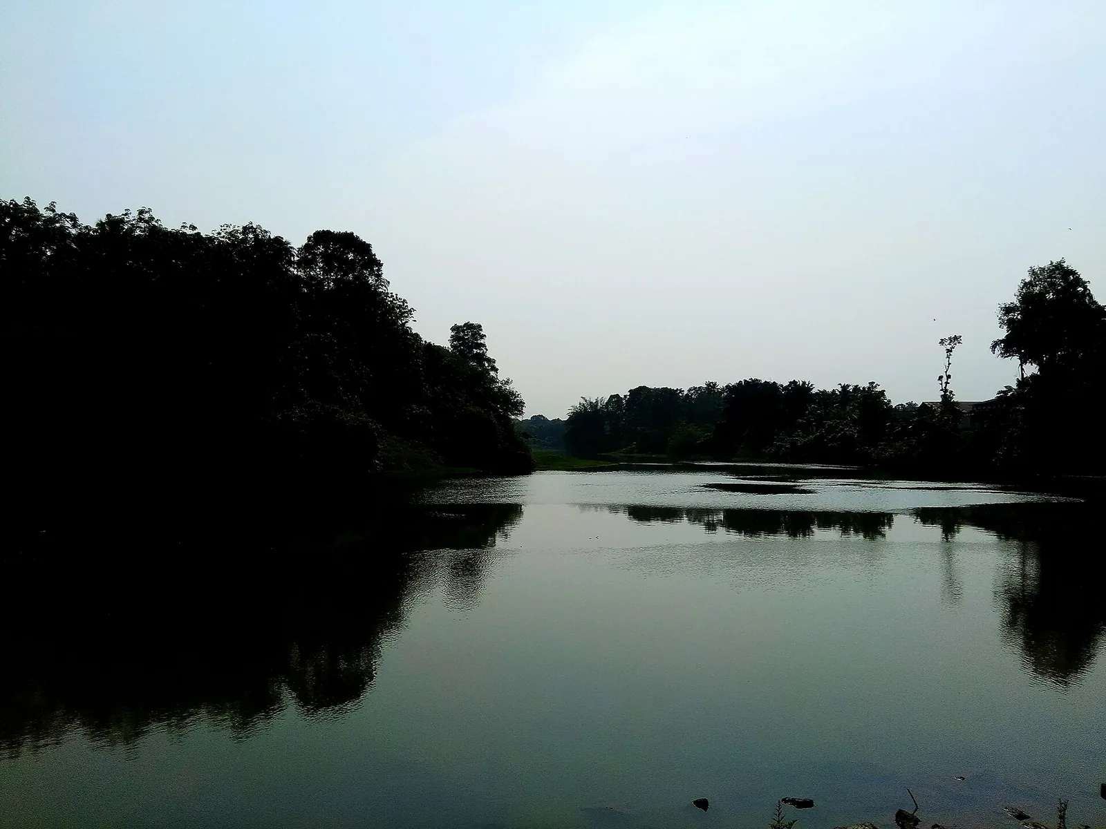 The Kallada river at Puthoor in flat grey light, a broad still reach reflecting dark tree-lined banks, with low hills on the horizon.