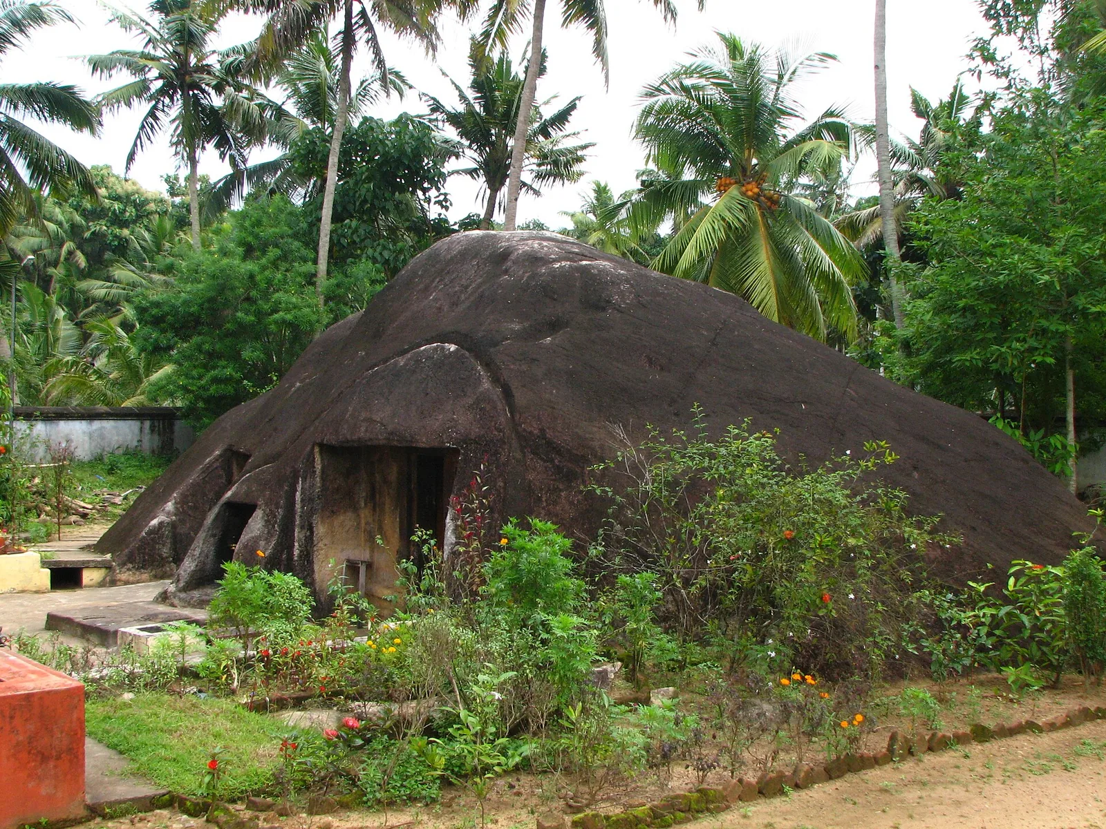 A large rounded granite boulder rising from a garden compound among coconut palms, with two door-sized openings of visibly different sizes cut into its lower face.
