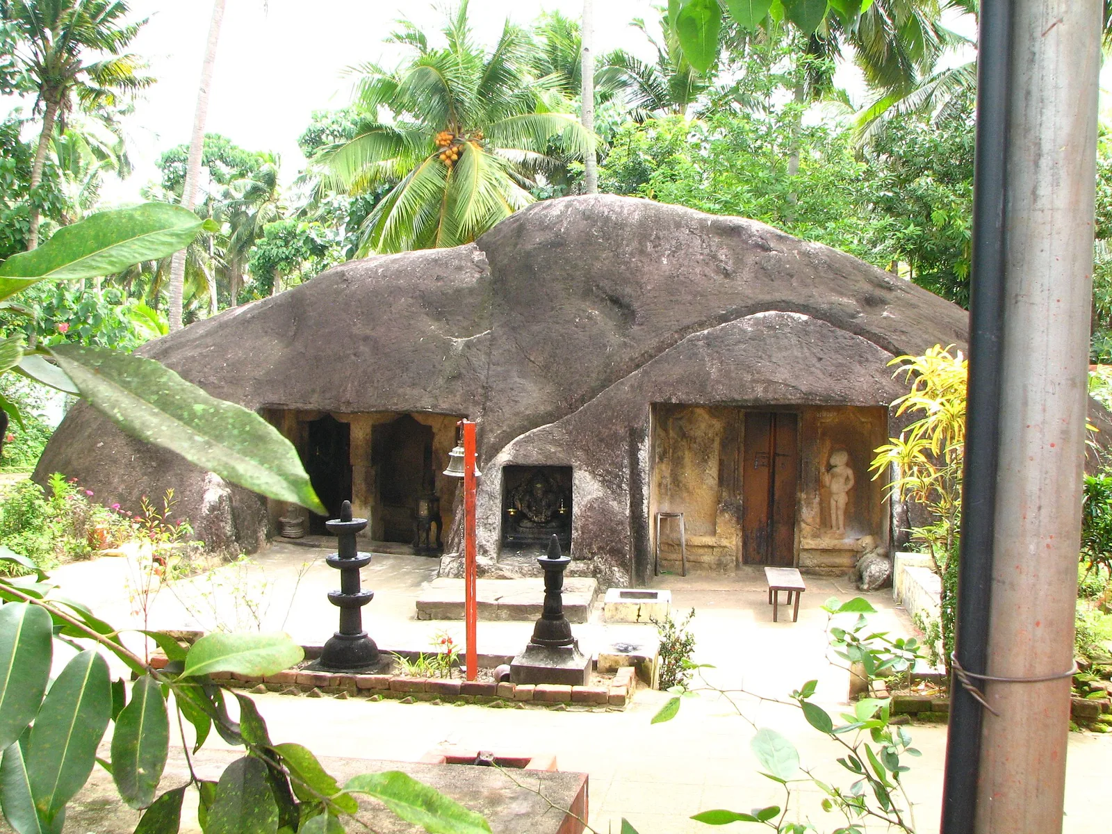 The Kottukal boulder seen across a swept compound with two cast-iron lamp-stands, a bell on a red post, and the Ganapathy visible in a low alcove between the two cave mouths.