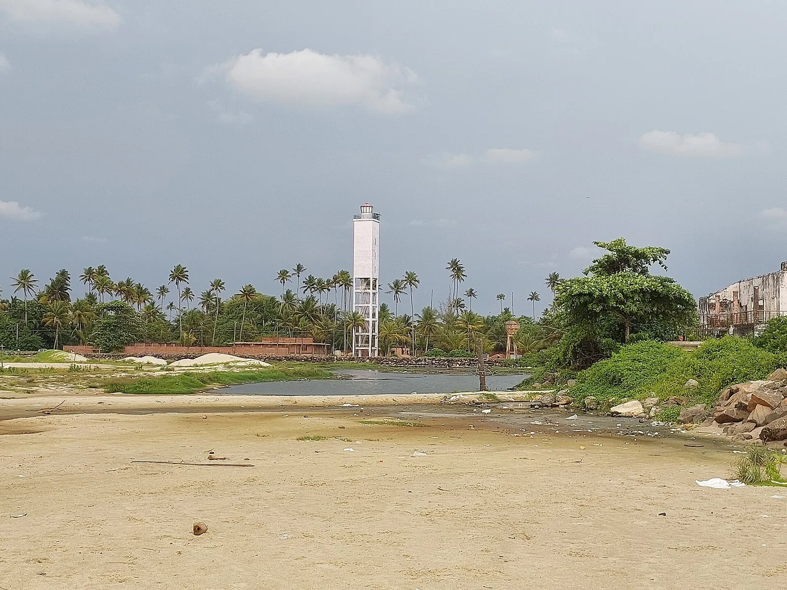 A white square lighthouse tower on an open four-column base seen across a shallow channel and a wide flat of pale sand, with coconut palms behind and rock revetment at right