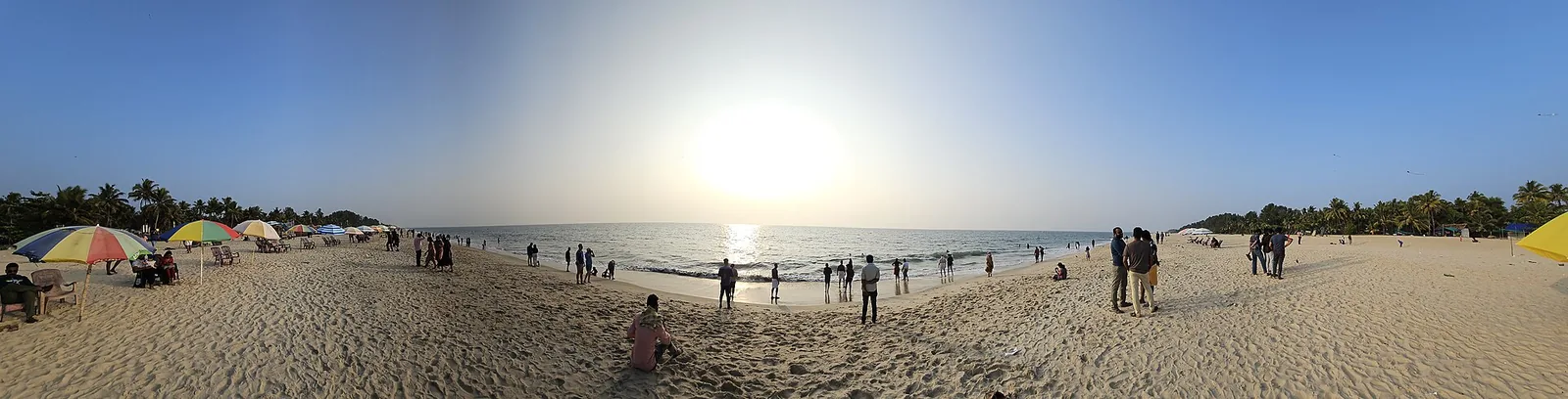 A wide panorama of Marari Beach at sunset, with coloured umbrellas and chairs at the left, dozens of people spread along the sand and standing in the shallows, and coconut palms behind on both sides