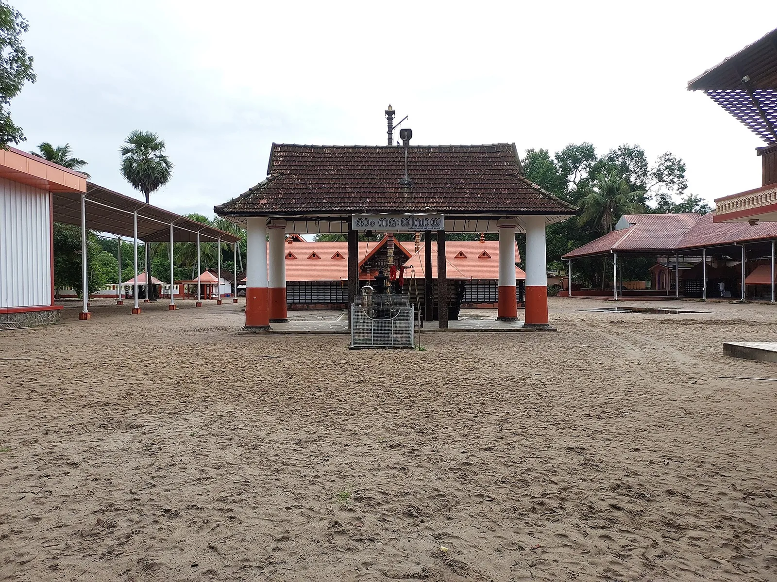 The sand courtyard of the Mararikulam Mahadeva temple, with a tiled namaskara mandapam, orange and white pillars and a board reading Om Namah Shivaya