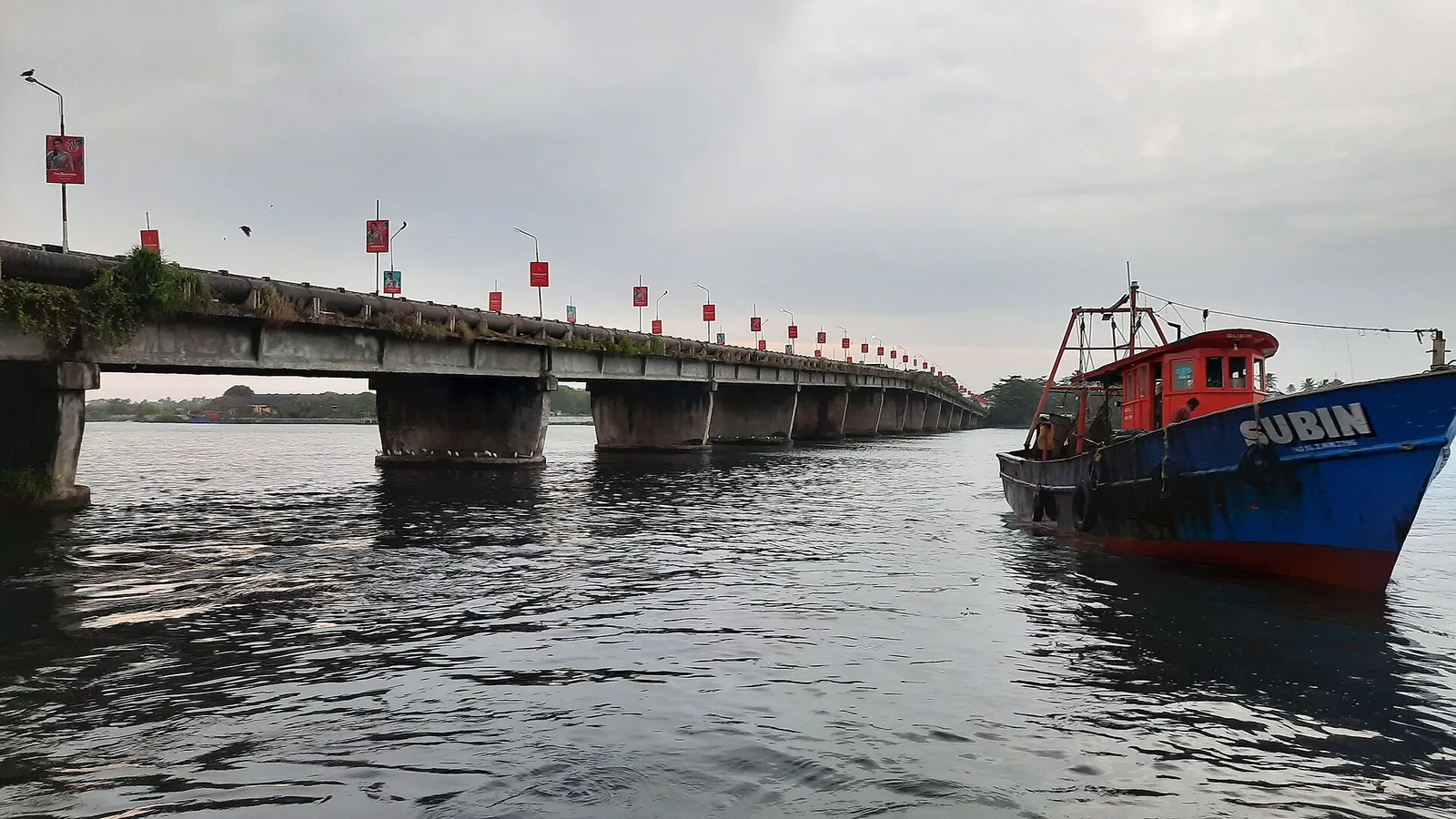 The low 1972 Neendakara road bridge on squat piers at water level, a large pipeline running along its flank and red banners on its lamp posts, with a blue-and-red trawler named SUBIN moored in the right foreground.