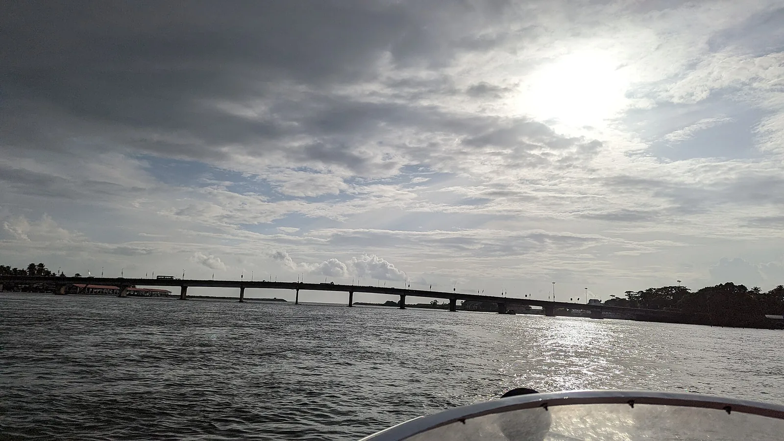 The high-level NH-66 bridge over the mouth of Ashtamudi Lake at Neendakara, seen low from a boat on the water, its long deck carried on tall piers with the auction hall visible at the far left of the shore.