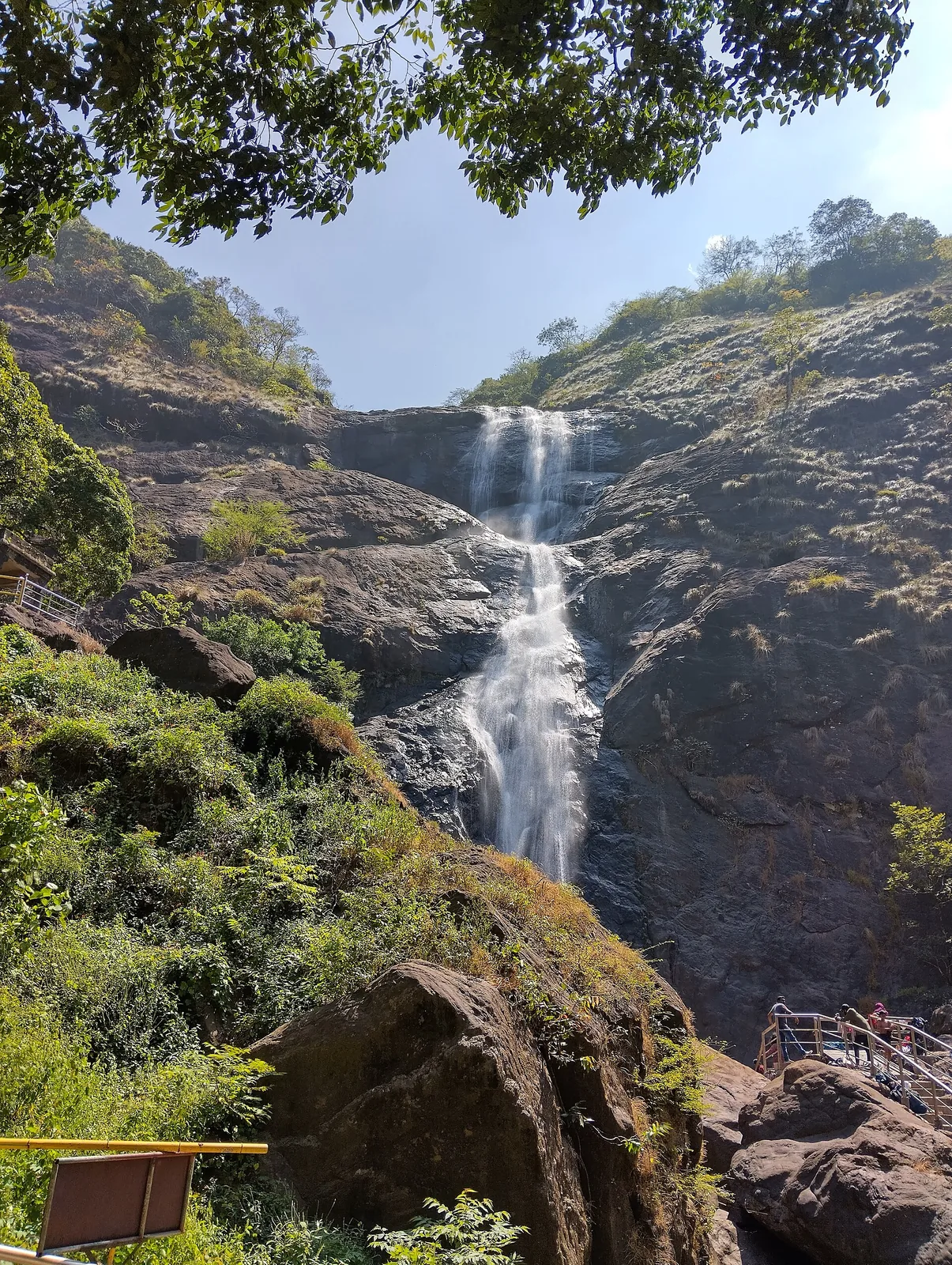 Palaruvi in low flow under a bright sky: a thin white ribbon comes over a high lip, slides down a broad grey granite apron and drops through a second cascade, with a yellow-railed metal viewing platform and visitors at lower right.