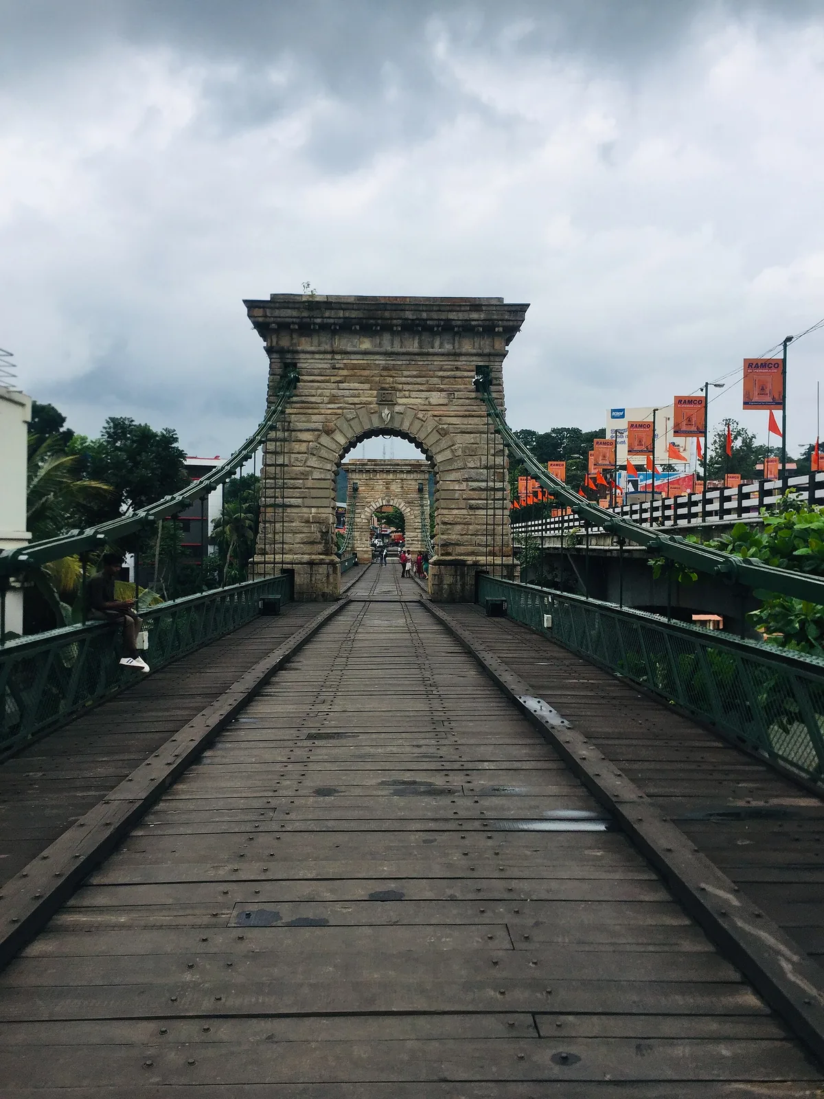 Looking down the timber deck of the Punalur suspension bridge under a storm sky, with two raised timber runners running the length of the planking towards the masonry arch.