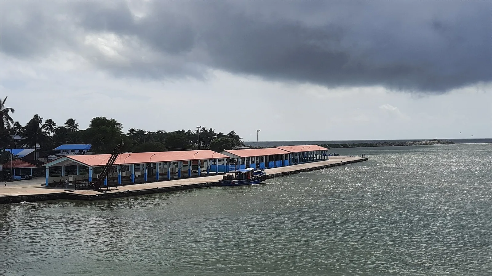 The long red-roofed open-sided auction hall on blue pillars along the Sakthikulangara quay, an ice-conveyor gantry at the near end and the breakwater running out to sea behind it with a light on its head, under a heavy grey storm sky, with a single small blue boat alongside an otherwise empty quay.