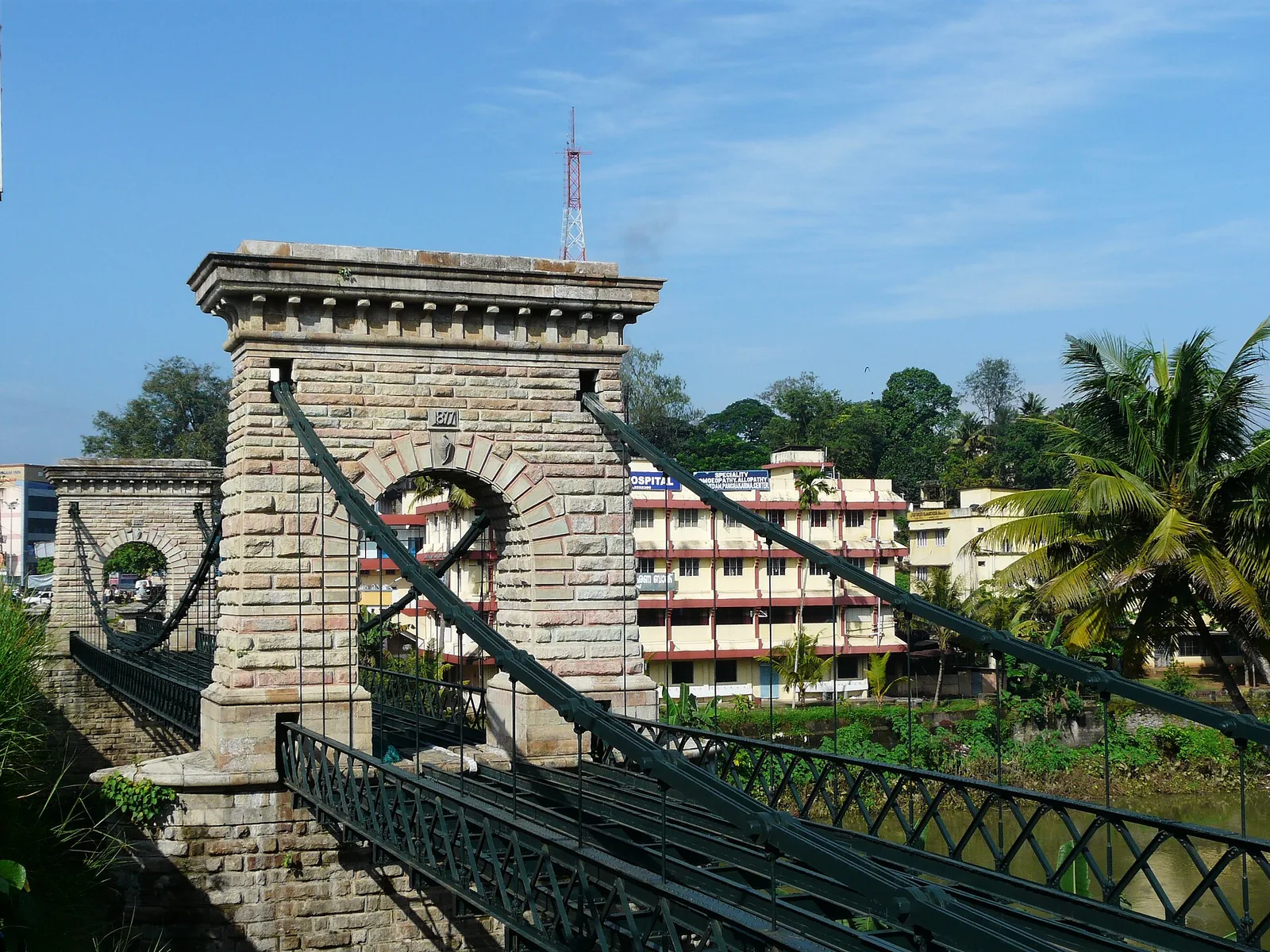 The Punalur suspension bridge seen from the bank, its iron chains sweeping over two masonry arch towers above the Kallada river.