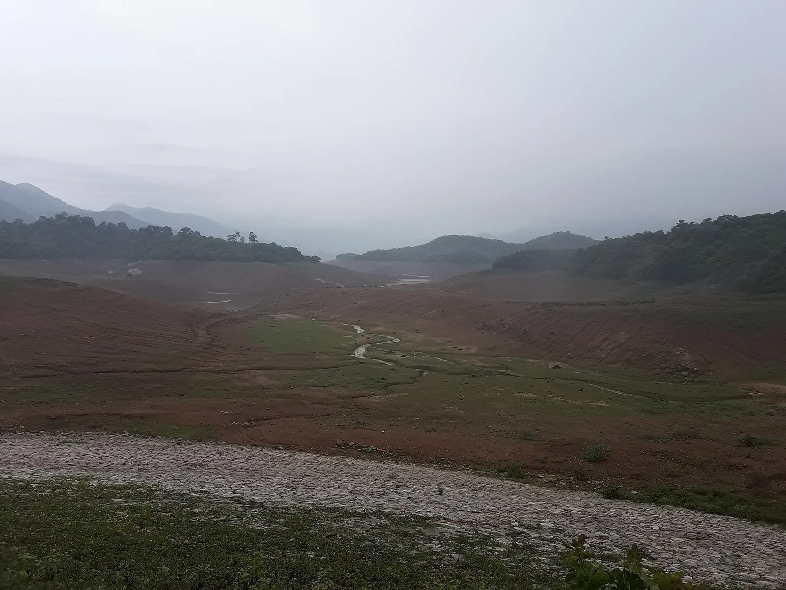 A wide reservoir basin drawn down to bare red earth, with a narrow stream channel winding through green flats on the valley floor and forested ridges disappearing into low monsoon cloud beyond.