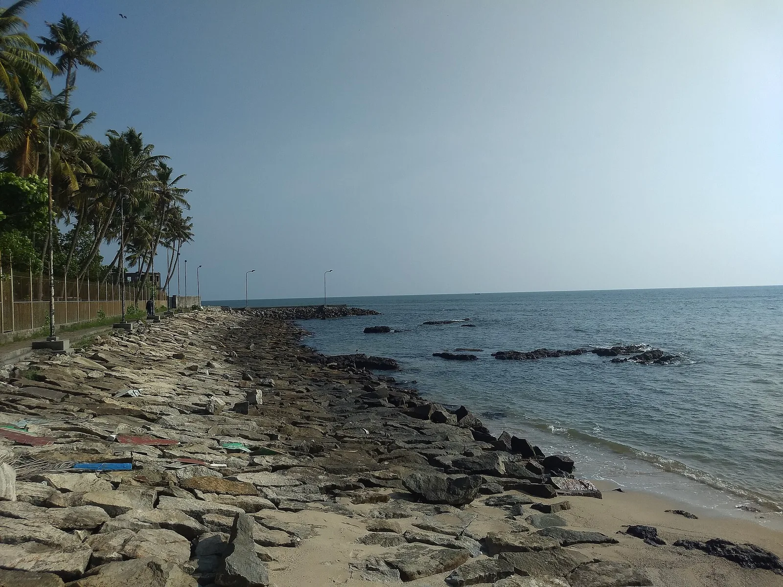 The shore at Thirumullavaram: a rock revetment along the waterline, a pocket of sand beyond, coconut palms behind, and open sea to the left.