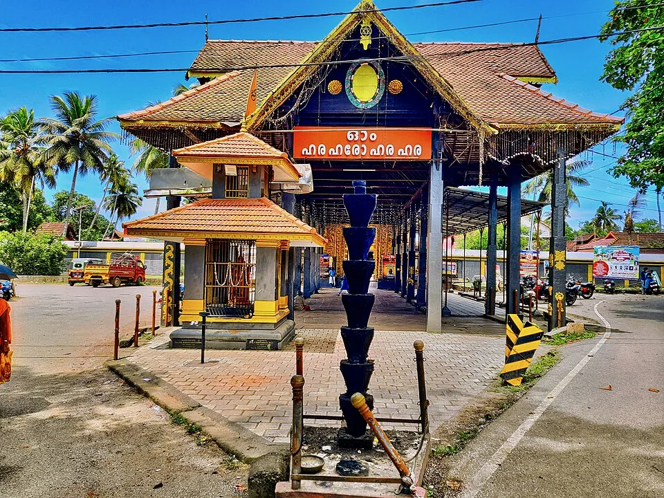 The street-front entrance of the Haripad temple with tiled roofs, an orange Malayalam board and a tall tiered black stone lamp pillar in the foreground