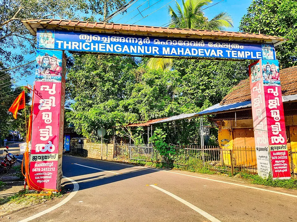 A tiled roadside arch across a road at Chengannur, its blue signboard reading “Thrichengannur Mahadevar Temple” in Malayalam and English, with red sponsorship panels down both piers.