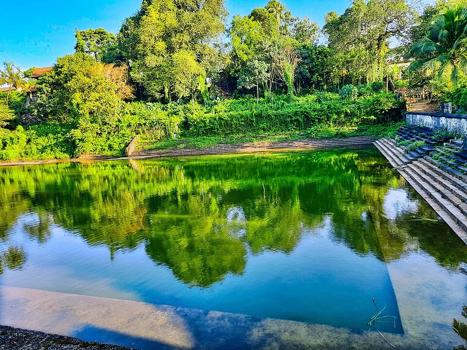 A rectangular temple tank at Chengannur with cut-stone ghat steps down one side, the water green with the reflection of trees.