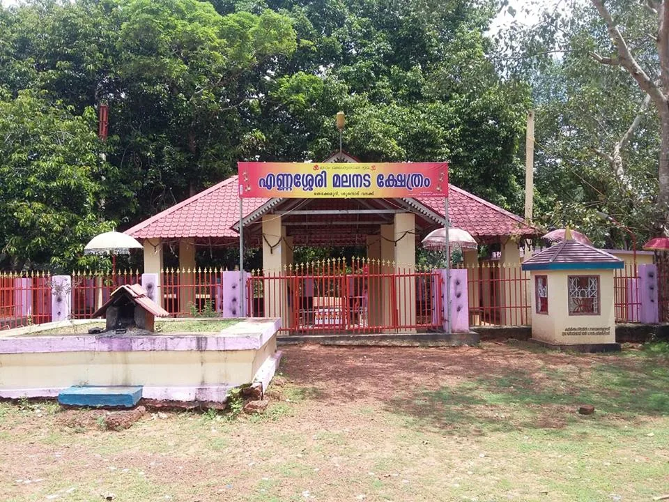 A small open pavilion under a red-tiled pyramidal roof behind a red painted railing, with a banner in Malayalam reading Ennasheri Malanada Kshethram, a low raised platform to the left and ceremonial umbrellas at the corners
