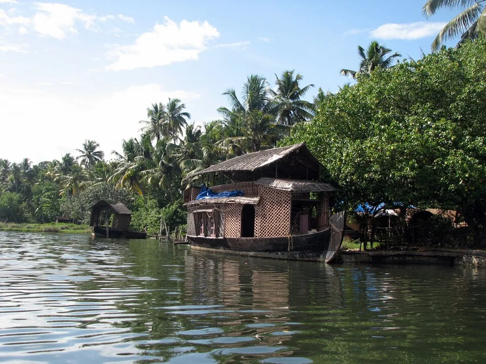 A thatched kettuvallam with woven cane walls moored against a green bank on a backwater canal, palms behind it