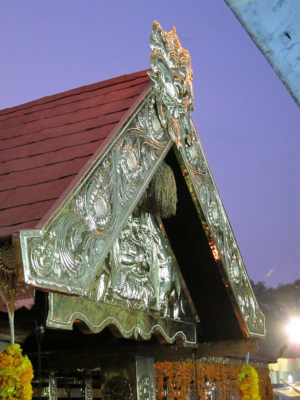 Close view of the temple gable at Kottankulangara: a triangular gable end entirely clad in beaten repoussé metal worked into scrollwork and figures, with red roof tiles running away behind it against a dusk sky