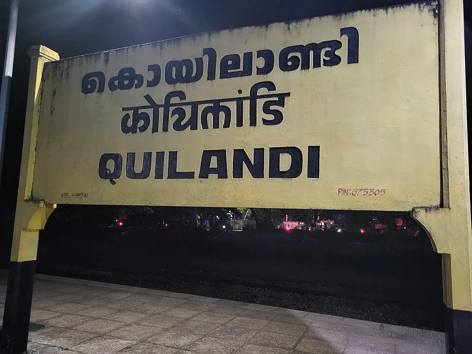 The Koyilandy railway station nameboard lit at night: a yellow-framed board with the town’s name painted in Malayalam and Hindi above the roman spelling QUILANDI, and the PIN code 673305 in the lower right corner.