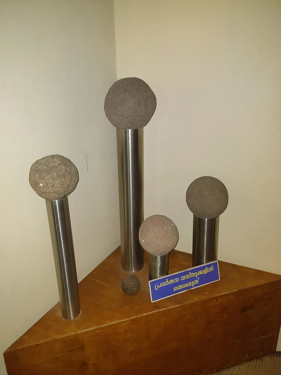 Five stone cannonballs of graduated size mounted on polished steel rods of different heights on a plain wooden plinth in a corner of the Kunjali Marakkar Museum, with a small loose stone ball beside them and a blue-and-white Malayalam sign reading “do not touch the exhibits”.