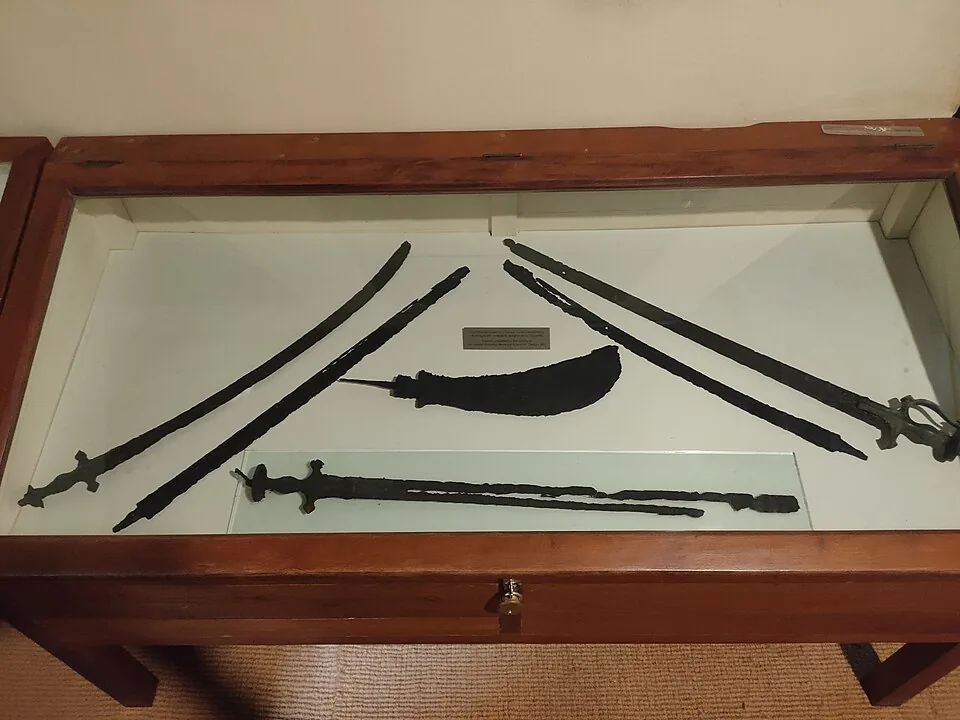 A flat wooden display case at the Kunjali Marakkar Museum holding six heavily corroded straight iron blades laid out in a fan, some cross-hilted and one with a knuckle guard, with a short curved weapon in a decayed scabbard across the middle.