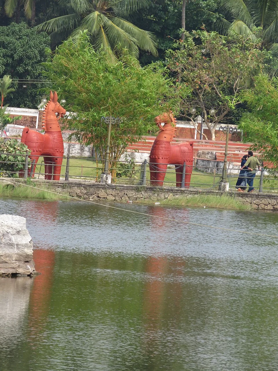 Two large red terracotta horses standing on the bank above the quarry lake at Sargaalaya, Iringal, with visitors walking the path behind them