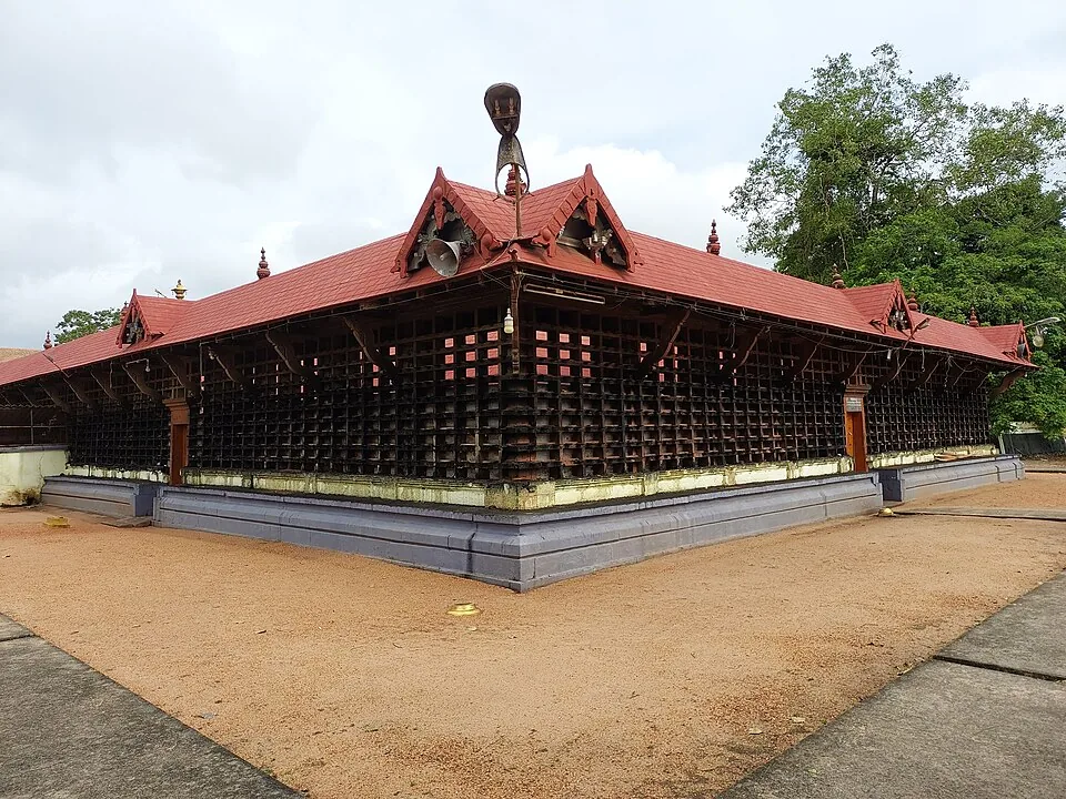 The outer wall of a Kerala temple covered corner to corner in a dense grid of small black oil-lamp niches, under a red tiled roof with bells hanging beneath the gables.